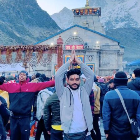 Devotee offering prayers outside Kedarnath Temple surrounded by pilgrims and snow-covered Himalayan mountains in Uttarakhand.