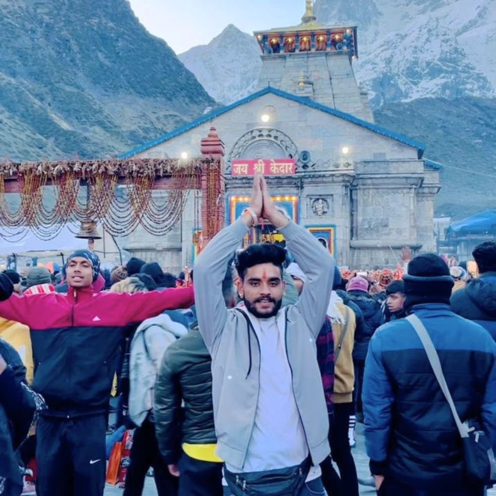 Devotee offering prayers outside Kedarnath Temple surrounded by pilgrims and snow-covered Himalayan mountains in Uttarakhand.
