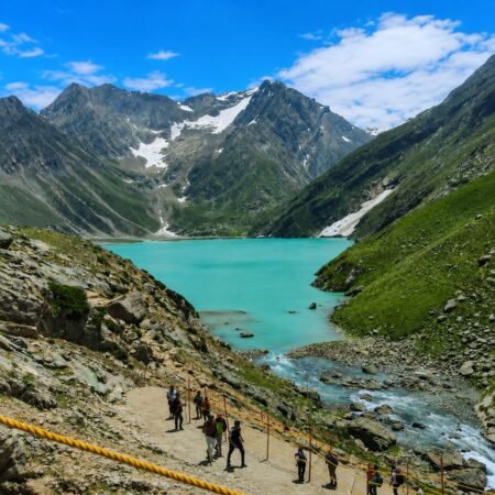 Pilgrims trekking along the Amarnath Yatra route beside a turquoise alpine lake surrounded by snow-capped Himalayan mountains in Jammu and Kashmir.