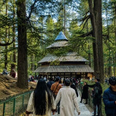 Front view of Hadimba Devi Temple surrounded by tall deodar trees in Manali, Himachal Pradesh, with visitors walking towards the historic wooden temple.