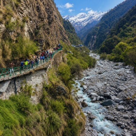 Panoramic view of the Yamunotri trek route near Janki Chatti featuring a cliffside walking trail, rocky Yamuna River valley, lush Himalayan forests, and snow-capped mountains in Uttarakhand.